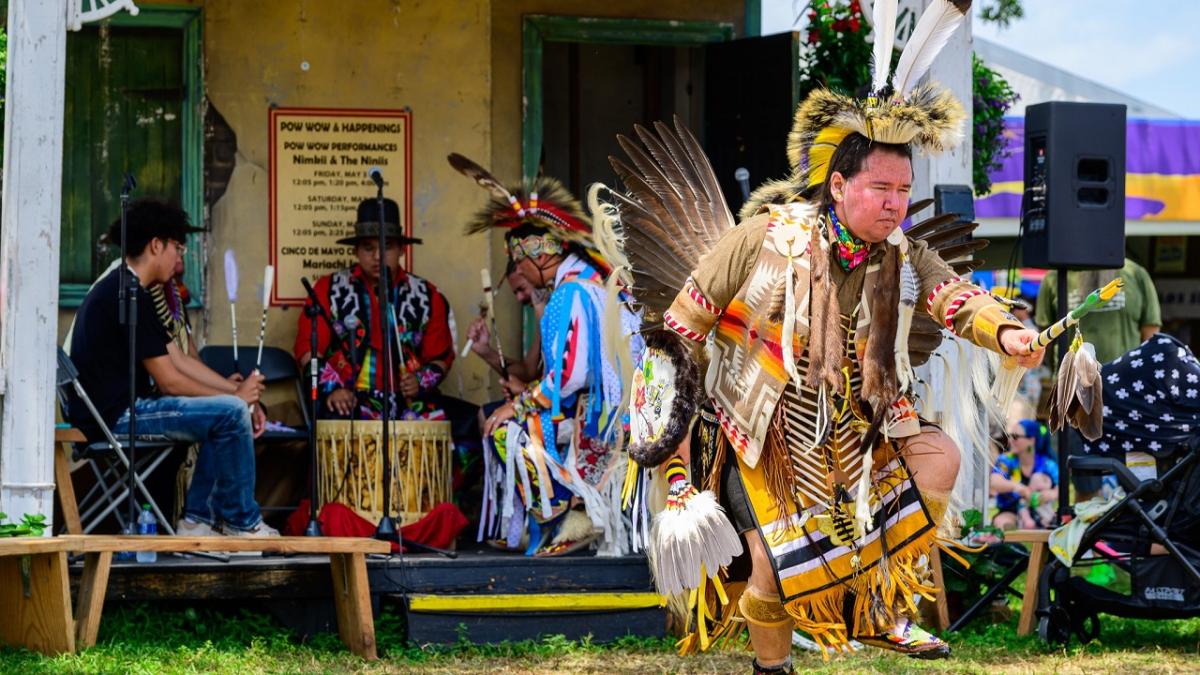 A person in ceremonial garments dancing, three behind them playing a drum.