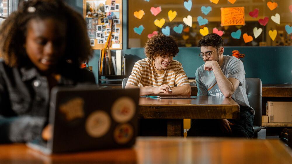 People seated at a table, looking at the same tablet. A person blurred in the foreground using a laptop. Colorful hearts on a window in the background.