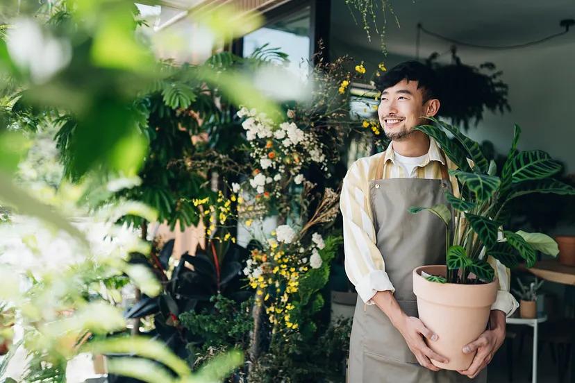 A smiling person holding a potted plant. Other plants hung around them.