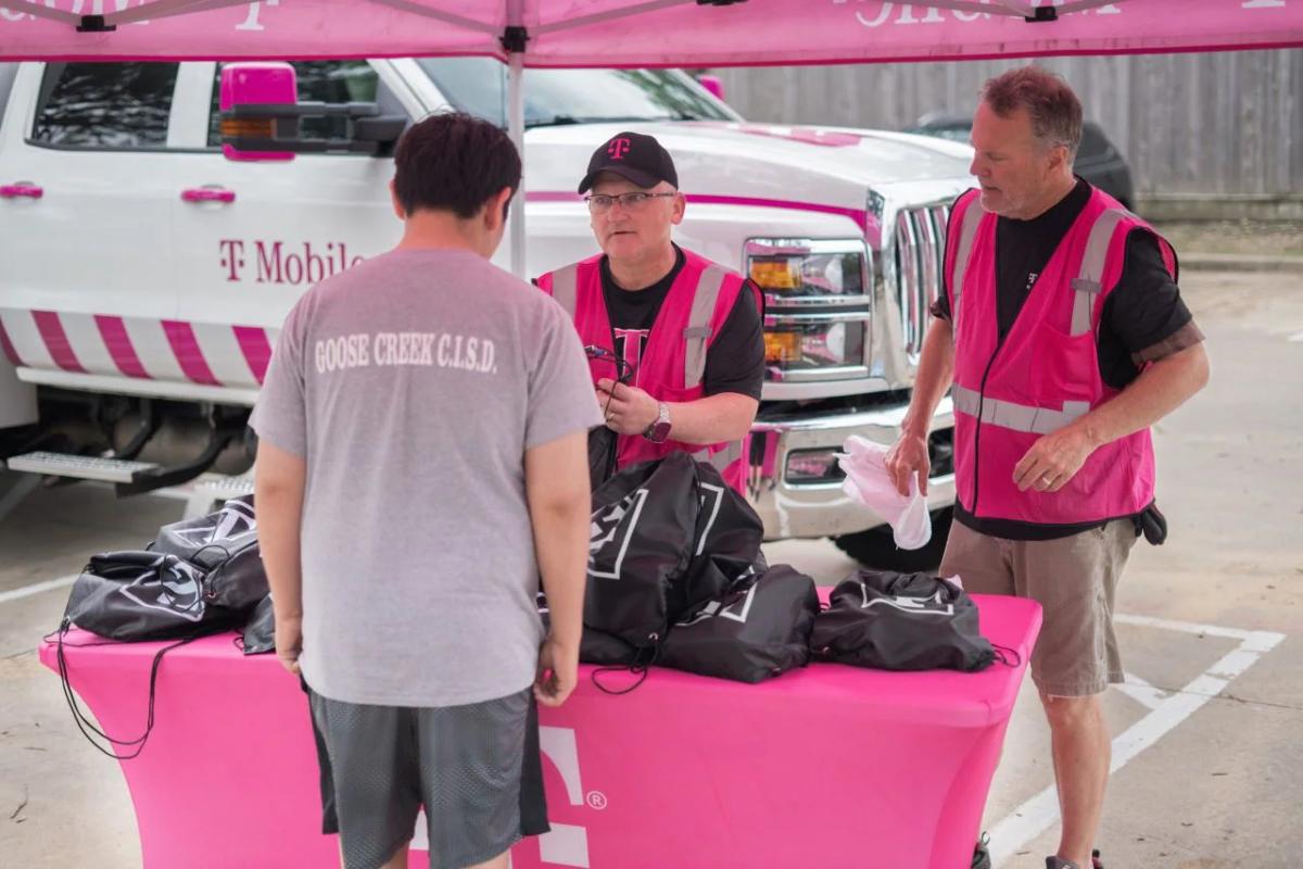 Two people in pink high-vis vests talk to another, a table full of black bags between them. 