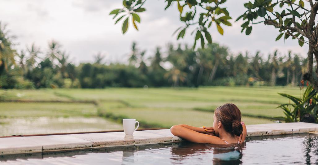 A person in an outdoor pool, arms crossed over the ledge overlooking a field and trees.