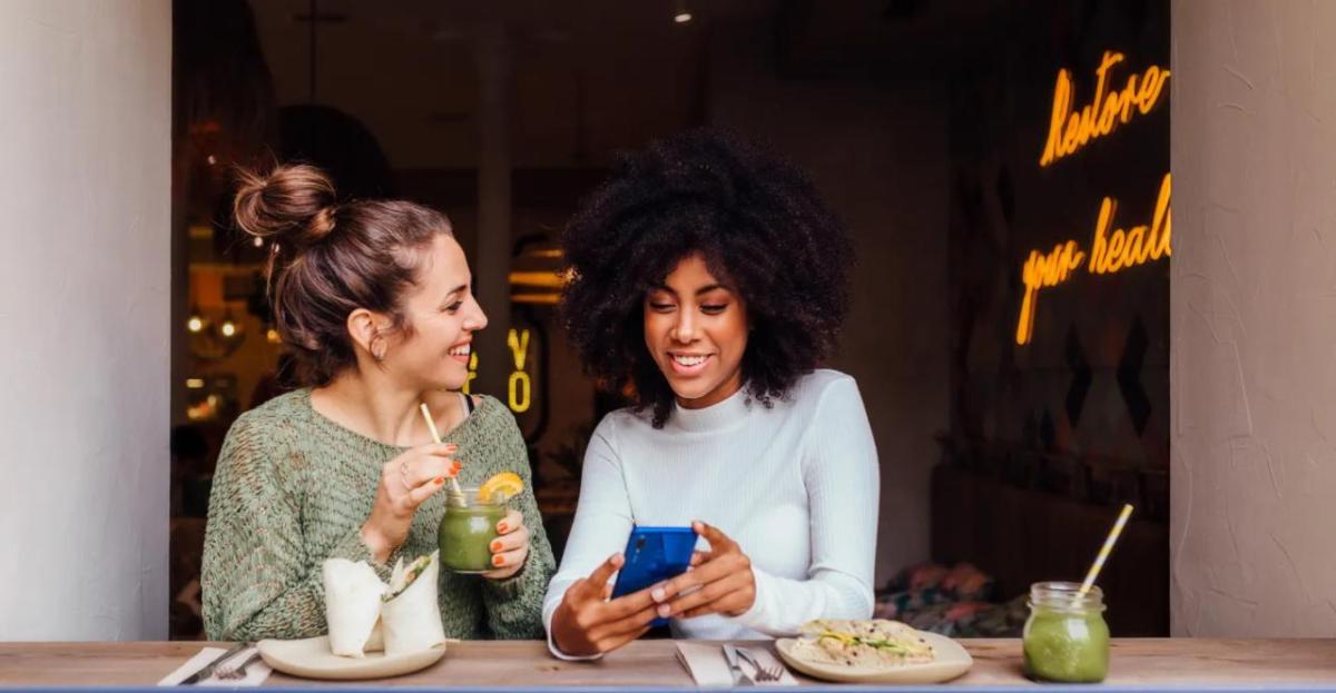 Two smiling people seated with food and drinks. One showing the other their phone.