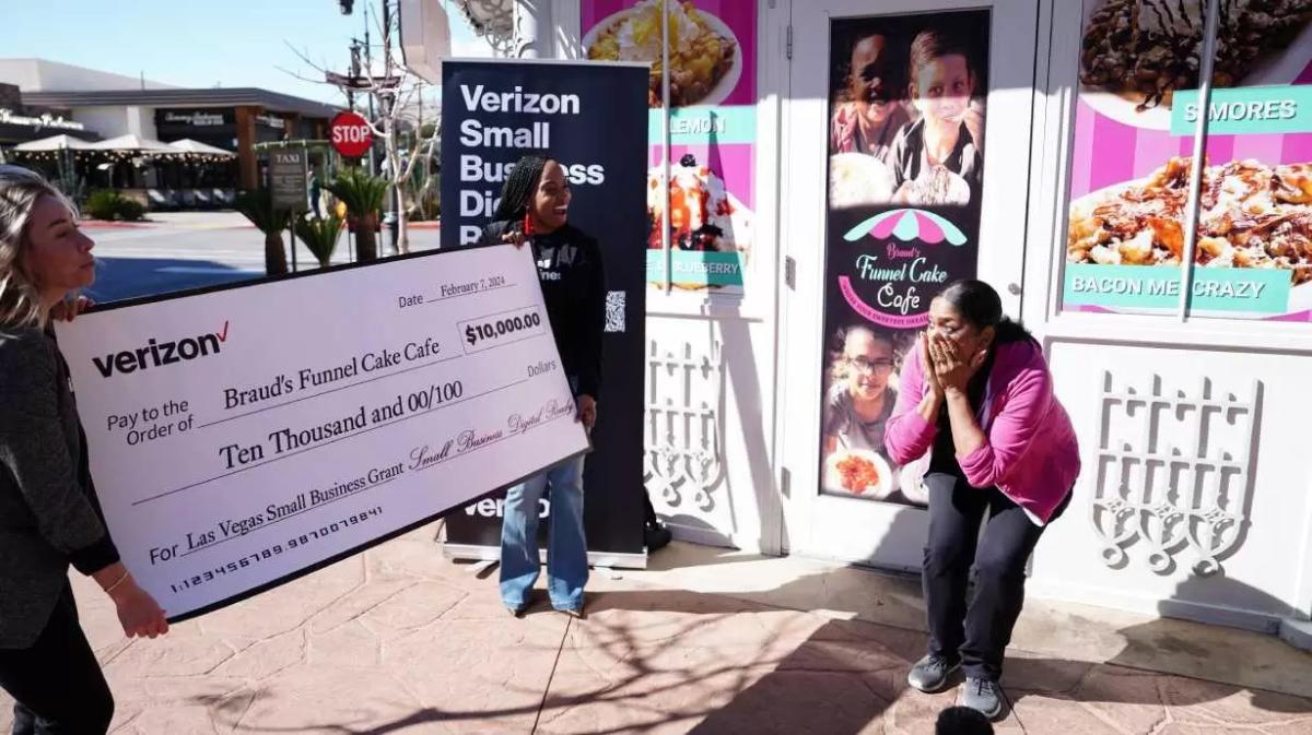 A surprised person covering their mouth as two other present a large check outside a bakery.