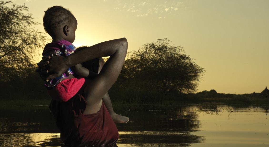 A mother in South Sudan wades through flood waters with her small child. Many refugees flee Sudan and seek shelter in South Sudan, which is burdened with its own climate shocks. / Photo by Peter Caton