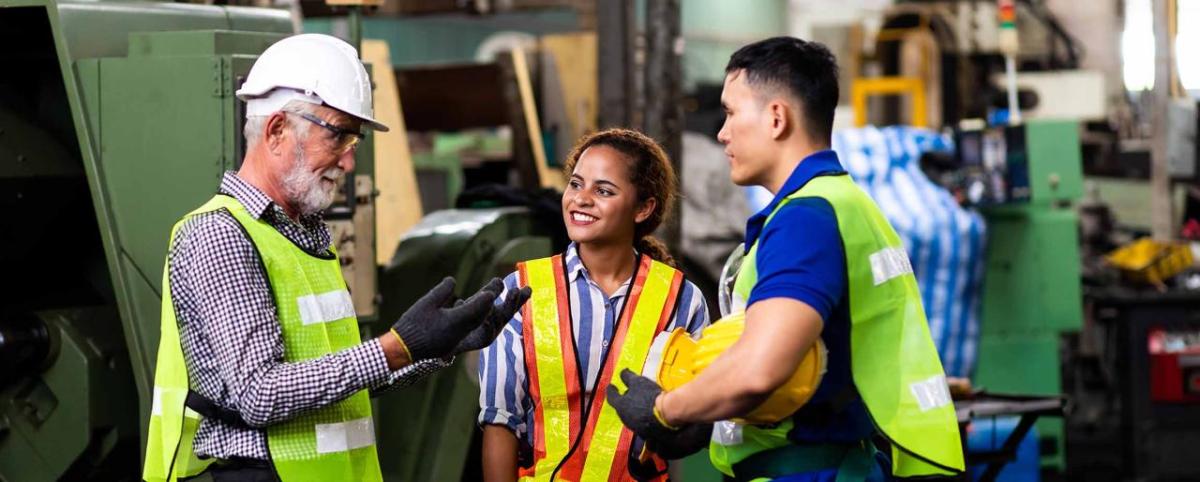 Two workers watch as a third talks with them. In an industrial setting, all are wearing high-vis vests.