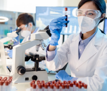 A person holding up a vial of blood next to a microscope