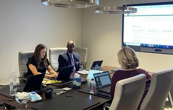 Three people seated at a conference table with laptops open. A digital display at the front.