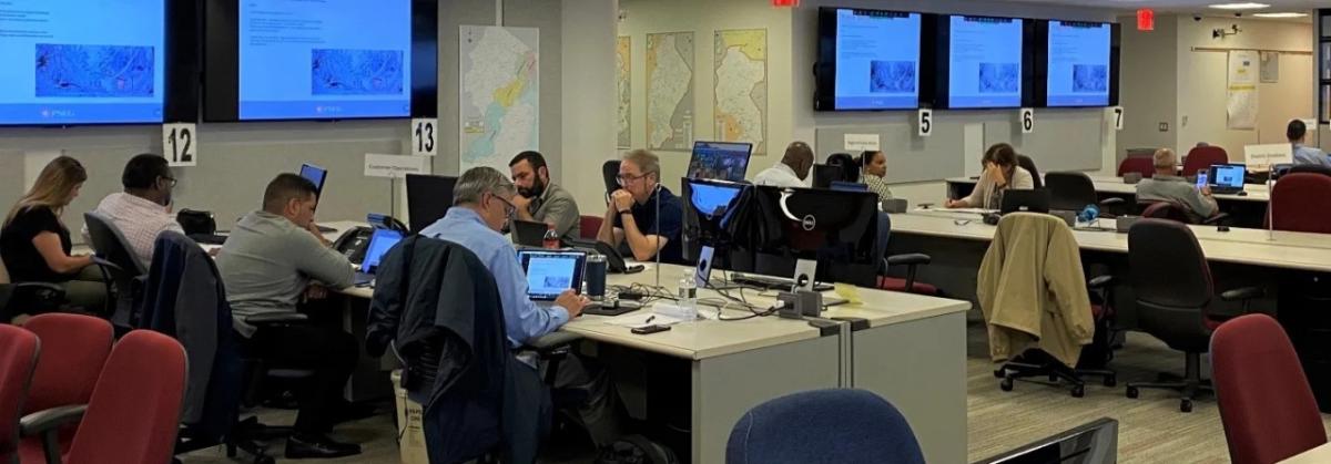 a large room with people seated at stationed tables, many screens on the wall