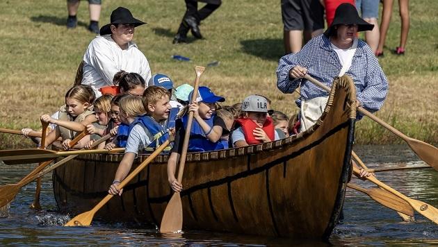 Children and two adults rowing in a large wooden canoe on a body of water near the shore.