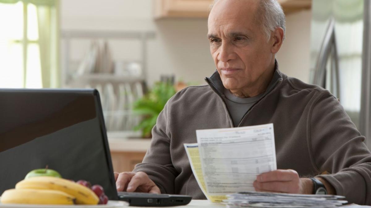 A person seated in front of a laptop computer in a home setting, holding papers in one hand.