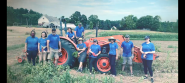 A group of people standing next to or on a large farm tractor in a field of growing crops. A home behind them. They're all wearing a blue shirt with "Alkermes" on it.